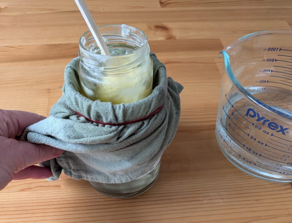 A jar held in a larger jar full of water, using a towel and rubber band.